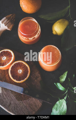 Flachbild-lay von zwei Gläser frisch gepressten Orange Saft oder Smoothie und frische Früchte auf Holzbrett über schwarzen Hintergrund, Ansicht von oben. Gesund l Stockfoto