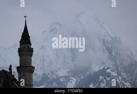 06. Mai 2019, Bayern, Schwangau: ein Turm von Schloss Neuschwanstein steht vor bewölkt und schneebedeckten Berge. Foto: Karl-Josef Hildenbrand/dpa Stockfoto