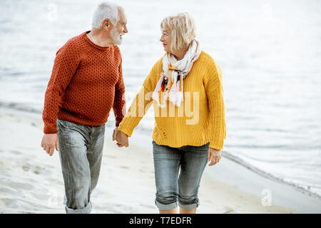 Schönes älteres Ehepaar in bunten Pullover zu Fuß am Sandstrand, genießen freie Zeit im Ruhestand in der Nähe des Meeres gekleidet Stockfoto