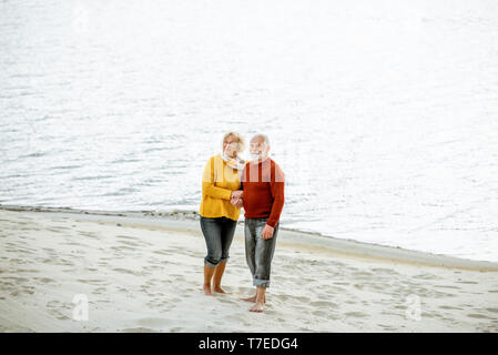 Schönes älteres Ehepaar in bunten Pullover umarmen am Sandstrand, genießen freie Zeit im Ruhestand in der Nähe des Meeres gekleidet Stockfoto