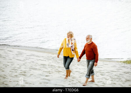 Schönes älteres Ehepaar in bunten Pullover zu Fuß am Sandstrand, genießen freie Zeit im Ruhestand in der Nähe des Meeres gekleidet Stockfoto