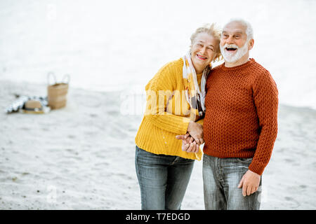 Portrait Of Happy senior Paar in bunten Pullover umarmen am Sandstrand, genießen freie Zeit im Ruhestand in der Nähe des Meeres gekleidet Stockfoto