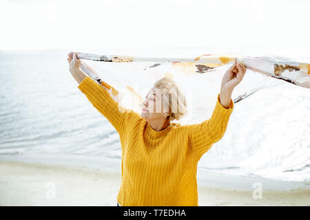 Ältere Frau im hellen Pullover genießen, Sea Breeze, holding Schal über dem Kopf am Sandstrand Stockfoto