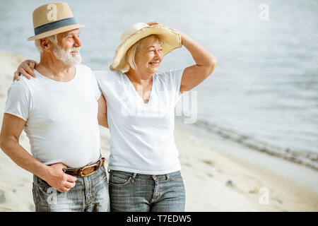 Gerne älteres Paar gekleidet in den weißen T-Shirts und Mützen zusammen zu Fuß am Sandstrand im Ruhestand Stockfoto