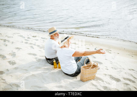 Senior Paar zusammen sitzen mit Getränke und Beutel auf dem Sandstrand, ihren Ruhestand in der Nähe des Meeres genießen, Rückansicht Stockfoto