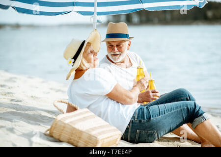 Gerne älteres Paar entspannende, liegend mit Getränken am Sandstrand, ihren Ruhestand in der Nähe des Meeres genießen. Stockfoto