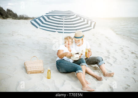 Gerne älteres Paar entspannende, zusammen liegen unter einen Sonnenschirm am Sandstrand, ihren Ruhestand in der Nähe des Meeres genießen.