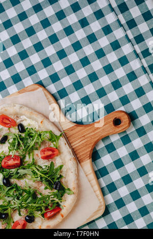 Neapolitanische Pizza auf Sahnesoße mit Thunfisch, Käse, Rucola, Basilikum, Tomaten, Oliven, bestreut mit Käse auf einem Holztisch auf einer Tischdecke in einer Zelle Stockfoto