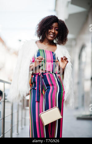 Modische african american woman in pink gestreifte Jumpsuit mit flauschigen Fellimitat Mantel stellte auf der Straße der Megapolis. Stockfoto