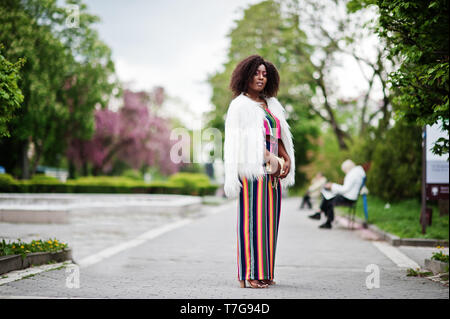 Modische african american woman in pink gestreifte Jumpsuit mit flauschigen Fellimitat Mantel stellte am Spring Bloom Street. Stockfoto