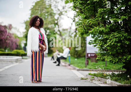 Modische african american woman in pink gestreifte Jumpsuit mit flauschigen Fellimitat Mantel stellte am Spring Bloom Street. Stockfoto