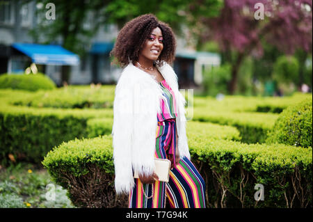 Modische african american woman in pink gestreifte Jumpsuit mit flauschigen Fellimitat Mantel stellte am Spring Bloom Street. Stockfoto