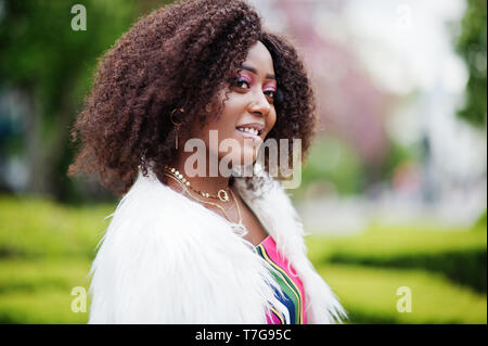 Modische african american woman in pink gestreifte Jumpsuit mit flauschigen Fellimitat Mantel stellte am Spring Bloom Street. Stockfoto