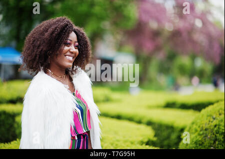 Modische african american woman in pink gestreifte Jumpsuit mit flauschigen Fellimitat Mantel stellte am Spring Bloom Street. Stockfoto