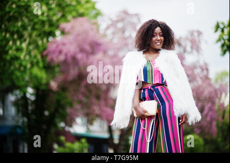 Modische african american woman in pink gestreifte Jumpsuit mit flauschigen Fellimitat Mantel stellte am Spring Bloom Street. Stockfoto