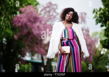 Modische african american woman in pink gestreifte Jumpsuit mit flauschigen Fellimitat Mantel stellte am Spring Bloom Street. Stockfoto