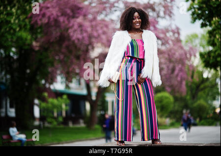 Modische african american woman in pink gestreifte Jumpsuit mit flauschigen Fellimitat Mantel stellte am Spring Bloom Street. Stockfoto