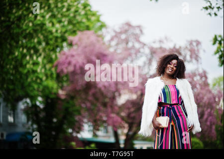 Modische african american woman in pink gestreifte Jumpsuit mit flauschigen Fellimitat Mantel stellte am Spring Bloom Street. Stockfoto