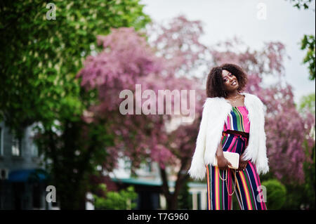 Modische african american woman in pink gestreifte Jumpsuit mit flauschigen Fellimitat Mantel stellte am Spring Bloom Street. Stockfoto