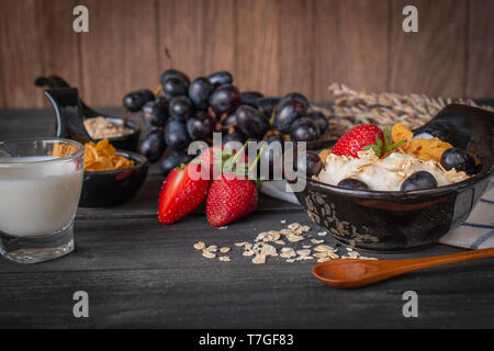 Joghurt mix Haferflocken, Erdbeeren und Trauben Richtfest in schwarz Schüssel auf Holz Tisch mit blau und weiß gestreifte Stoff, Löffel, Erdbeere, Milch in Glas, Gr Stockfoto