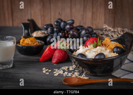 Joghurt mix Haferflocken, Erdbeeren und Trauben Richtfest in schwarz Schüssel auf Holz Tisch mit blau und weiß gestreifte Stoff, Löffel, Erdbeere, Milch in Glas, Gr Stockfoto