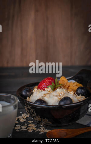 Joghurt mix Haferflocken, Erdbeeren und Trauben Richtfest in schwarz Schüssel auf Holz Tisch mit blau und weiß gestreifte Stoff, Löffel, Haferflocken und Milch in Glas pla Stockfoto