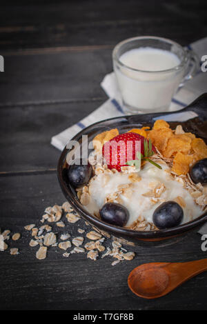 Joghurt mix Haferflocken, Erdbeeren und Trauben Richtfest in schwarz Schüssel auf Holz Tisch mit blau und weiß gestreifte Stoff, Löffel, Haferflocken und Milch in Glas pla Stockfoto