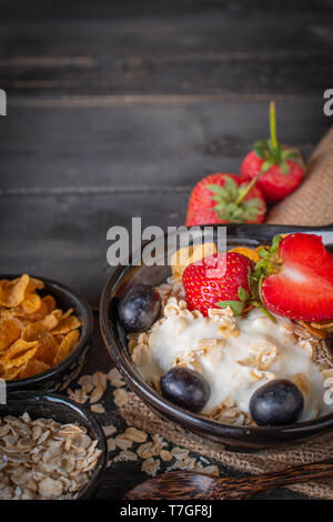 Joghurt mix Haferflocken, Erdbeeren und Trauben Richtfest in schwarz Schüssel auf Sack und Holz Tisch mit Löffel, Haferflocken, Cornflakes und Erdbeere platziert um. Stockfoto