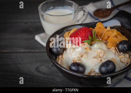 Joghurt mix Haferflocken, Erdbeeren und Trauben Richtfest in schwarz Schüssel auf Blau-weiß gestreifte Stoff und Holz Tisch mit Löffel und Milch im Glas gesetzt Ba Stockfoto