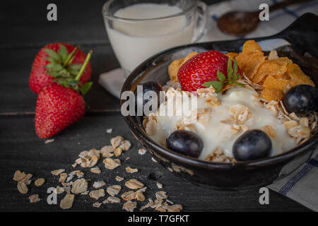 Joghurt mix Haferflocken, Erdbeeren und Trauben Richtfest in schwarz Schüssel auf Blau-weiß gestreifte Stoff und Holz Tisch mit Löffel, Milch in Glas, Erdbeere Stockfoto