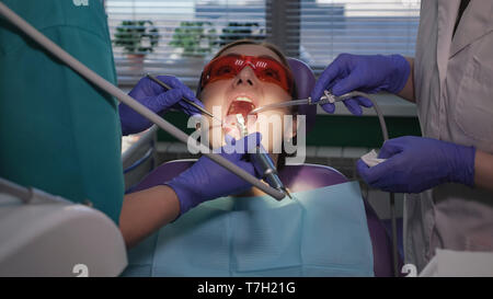 Zahnarzt ist die Betreuung von Patienten mit schwarzer Hohlraum mit elektrischen zahnmedizinischen Bürste. Krankenschwester mit Speichel Auswerfer in der Nähe der Patientin helfen, Doktor. Woman's Portrait in den zahnmedizinischen Stuhl. Stockfoto