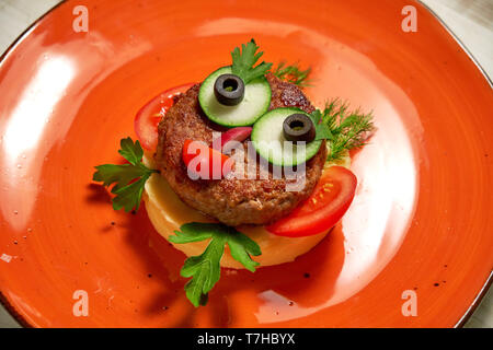 Das Restaurant serviert ein Gericht auf der Speisekarte des Kinder. Kartoffelpüree mit Schnitzel und Gemüse auf eine orange Gesicht in der Form eines Gesichts Stockfoto