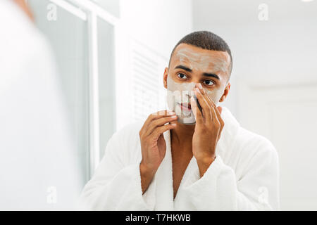 Junger Mann mit weißer Bademantel, die Gesichtsmaske im Badezimmer Stockfoto