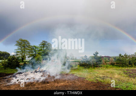 Rauch von einem großen ländlichen Lagerfeuer in der Landschaft langsam in die Wolken Himmel mit einem wunderschönen Regenbogen bei leichter Regen Stockfoto