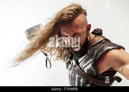 Gott des Donners. Blondes langes Haar und muskulös männliches Modell in der Viking leder Kostüm mit dem großen Hammer cosplaying Thor auf weißem studio Hintergrund isoliert. Fantasy Warrior, antiken Schlacht Konzept. Stockfoto