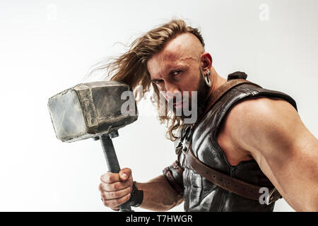 Gott des Donners. Blondes langes Haar und muskulös männliches Modell in der Viking leder Kostüm mit dem großen Hammer cosplaying Thor auf weißem studio Hintergrund isoliert. Fantasy Warrior, antiken Schlacht Konzept. Stockfoto