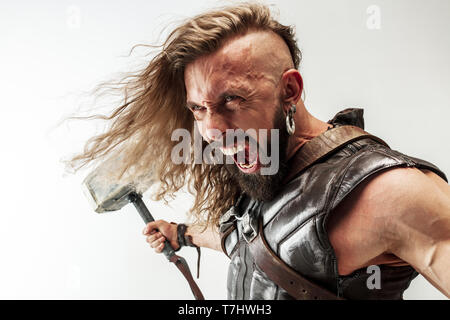 Gott des Donners. Blondes langes Haar und muskulös männliches Modell in der Viking leder Kostüm mit dem großen Hammer cosplaying Thor auf weißem studio Hintergrund isoliert. Fantasy Warrior, antiken Schlacht Konzept. Stockfoto