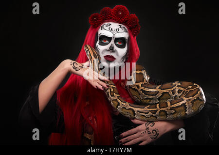 Spooky Frau mit Zucker frei machen, rote Haare, rote Augen und Lippen auf Kamera. Modell mit Stirnband von roten Blumen posing und halten grosse Schlange. Stockfoto