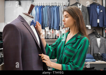 Bg Wahl der stilvolle Kleidung für Männer in der Boutique. Jacken in der Zeile hängend auf Kleiderbügeln. Schönen shop Berater stehen in der Nähe der Puppe im grauen Anzug, weißes Hemd, blau Bow Tie, Arbeiten mit Kleidung. Stockfoto