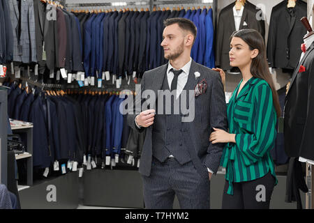 Stattliche Gentleman in der Trendfarbe Grau gestreiften Anzug und Krawatte schwarz, weißes Hemd im Store posiert, entfernt. Hübsches Mädchen Shop Assistant hilft Kunden bei der Auswahl der Kleidung. Stockfoto