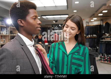 Riesige Auswahl Kleidung für Intelligenz Mann. Junger Mann, afrikanische in Boutique Wahl binden. Lächelnde Frau, Assistant hilft auf rot Krawatte versuchen, in der Nähe von Shirt des Mannes. Stockfoto