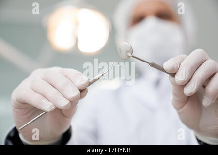 Stomatologist tragen in der medizinischen Maske, Kappe und weiße Uniform, Handschuhe bei Patienten suchen von unten. Zahnarzt Holding professionelle Restaurierung Instrumente in den Händen. Stockfoto