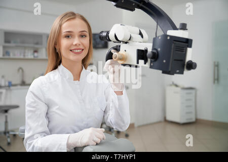 Gerne weiblichen Zahnarzt in der Zahnheilkunde Privatklinik posieren. Schöne und bezaubernde blonde Frau an der Kamera schauen und lächeln, Stomatologist Hand auf zahnmedizinische Mikroskop. Stockfoto