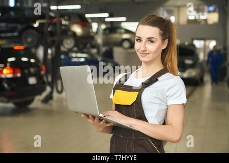 Schöne junge Frau arbeitet als Mechaniker holding Notebook in die Hände. Hübsches Mädchen mit Blick auf die Kamera oder Lächeln, posieren. Weibliche Arbeitnehmer tragen im weißen T-Shirt und braunen Overalls. Stockfoto