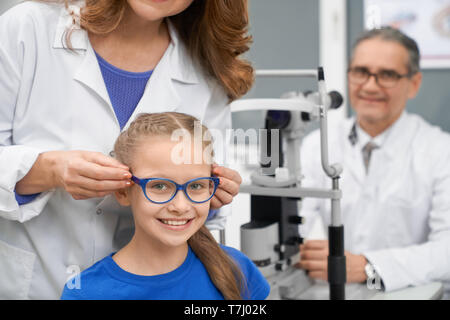 Arzt Augenarzt die geduldigen Sehvermögen. Medizinischer Arbeiter im weißen Mantel Wahl Gläser für Teenager. Mädchen in Blau Brille sitzen, lächelnd und mit Blick auf die Kamera. Augenheilkunde Kabinett. Stockfoto