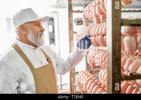 Wurst Fleisch Fabrik Arbeiter. Wurst in Reihe hängen. Ältere Arbeitnehmer, zu beobachten und zu prüfen, testen Würstchen. Mann in weißer Uniform, weiße Kappe, braun Schürze, Gummihandschuhe. Stockfoto