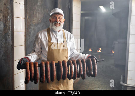 Geräucherter sassages in Reihe hängen. Stattlich Raucher in der räucherei mit geräucherter Wurst, an der Kamera schaut. Ältere Mann, der in der weißen uhiform, braune Schürze, lächelnd, posieren. Stockfoto