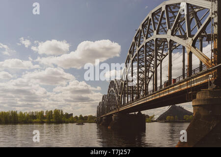 Eisenbahnbrücke über den Fluss Daugava in Riga mit einem Boot und der Lettischen Nationalbibliothek auf der Bank im Frühjahr Stockfoto