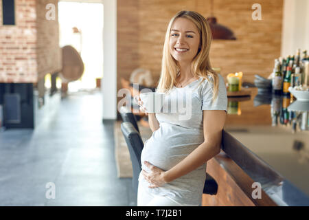 Lächelnd glückliche junge schwangere Frau entspannend lehnte sich gegen einen Tresen mit einer Tasse heißen Tee mit Ihrem Babybauch in ihrer Hand, mit der sie Anleihen mit h Stockfoto