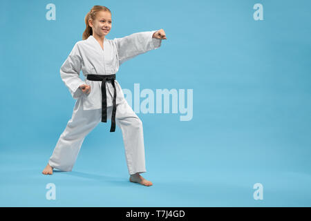 Ansicht von der Seite der fröhlichen Kämpferin in Haltung und Stanzen mit Hand im Studio. Lächelnde Mädchen tragen Kimonos Üben von Karate und Jiu-jitsu auf blauem Hintergrund isoliert. Konzept der Sport. Stockfoto